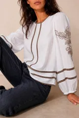 Blusa blanca de manga larga con detalles de bordados florales en tono grisáceo y bandas decorativas en el frente y mangas. Cuenta con cuello redondo y puños elásticos.