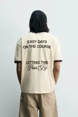 Camiseta de corte regular, color beige con cuello y bordes de mangas en color marrón oscuro. Presenta un diseño de texto bordado en el pecho con la frase 'Letting Time Pass By'.