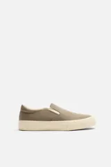 Zapatilla tipo slip-on de lona color taupe, con elástico lateral para facilitar el calce y suela de goma gruesa color crema.