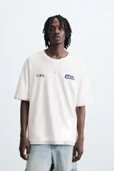 Camiseta de manga corta y cuello redondo, color blanco roto, con efecto de doble capa (la capa inferior gris asoma en el ruedo). Presenta estampados pequeños en azul marino en el pecho: un logo con texto 'Idle Eden' y otros símbolos.