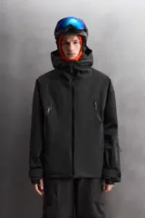Cazadora deportiva de esquí en color negro, confeccionada en tejido técnico impermeable de tres capas con costuras y cremalleras termoselladas. Cuenta con capucha ajustable, ventilación en axilas, puños con ajuste de tira adherente y puño interior elástico con abertura para el pulgar. Incluye sistema de rescate RECCO y faldón interior para protección contra el frío.