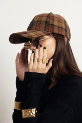 Gorra de estilo béisbol con estampado de cuadros tipo tartán en tonos marrones y grises. Presenta un parche frontal de cuero sintético con la marca grabada y cierre ajustable en la parte posterior.