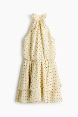 Vestido corto de gasa transparente en color beige con estampado de lunares negros. Presenta un diseño con escote halter, tiras anchas para atar en la nuca, espalda abierta, cintura elástica fruncida y falda escalonada con vuelo. Incluye forro interior.