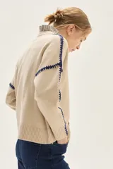 Sweater de tejido de punto color beige, con cuello redondo y detalles de bordado en zigzag en color azul oscuro a lo largo de las mangas y hombros. Presenta un pequeño bordado con texto en el pecho.