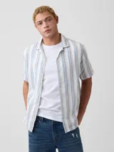 Camisa de manga corta de lino con diseño de rayas verticales en tonos blancos, azules y marrones, cuello camisero y cierre frontal de botones.