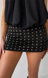 Vestido corto negro con escote halter drapeado y falda ajustada con detalle de tachas doradas cuadradas.