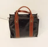 Cartera tipo bowling color beige con detalles en cuero negro, doble asa corta y cierre superior con cremallera.
