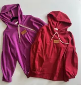 Canguro con capucha y cordones ajustables, confeccionado en tejido de punto. Presenta mangas largas abullonadas con puños elásticos y un parche de cuero sintético con logo en el pecho.