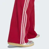 Pantalón deportivo Adidas Firebird Pinstripe color rojo con finas rayas verticales y las clásicas tres tiras blancas en los laterales. Cuenta con tiro medio, bolsillos laterales con cierre y el logo del trifolio bordado en blanco.