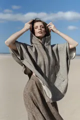 Capa amplia de lana fina sin teñir, con capucha, cierre frontal metálico y bolsillos delanteros. Presenta un diseño con canesú en la espalda y botones laterales que permiten ajustar la forma de las mangas.
