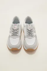 Zapatilla deportiva de diseño urbano, combinando paneles de tela blanca y gamuza sintética en tono gris claro. Cuenta con cordones grises, suela de plataforma con entresuela blanca y base dentada en color caramelo.