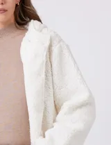 Campera tipo teddy de textura suave y esponjosa en color blanco, con cierre frontal mediante botones y detalles de apliques en cuero sintético negro en la solapa. Presenta bolsillos frontales y un diseño cómodo y abrigado.