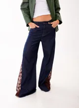 Pantalón de jean estilo wide leg en color azul oscuro, con paneles laterales de encaje en tono marrón rojizo. Presenta un corte amplio y estructura firme sin elasticidad.