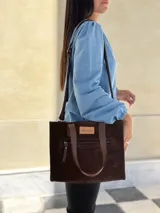 Cartera tipo tote de gamuza color chocolate, con doble asa de mano y correa larga ajustable y extraíble. Presenta un bolsillo frontal con cierre metálico y una etiqueta de cuero con el logo de la marca en el frente.
