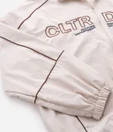 Campera de tejido técnico color beige con detalles de costuras en contraste color marrón. Presenta cuello alto, cierre frontal con cremallera, mangas largas con puños elásticos y estampa frontal con texto. Incluye un detalle bordado en la manga.