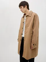 Campera tipo trench coat de corte recto y largo medio, color tostado. Presenta cuello camisero, cierre frontal con botones oscuros y puños ajustables con trabilla y botón.