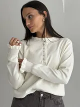 Sweater de punto color blanco, con cuello redondo y cierre frontal parcial con botones. Presenta mangas largas con puños acanalados y detalle de volados en los extremos.