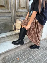Bota de caña alta estilo biker, confeccionada en cuero sintético negro. Presenta una suela track con plataforma, puntera redondeada y un detalle de arnés con hebilla metálica y tachas en la zona del tobillo.