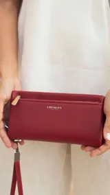 Billetera de mujer tipo sobre con correa de mano extraíble, confeccionada en material texturizado color tostado. Presenta un bolsillo frontal con cierre, compartimentos internos para tarjetas y logo de la marca grabado en el frente.