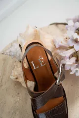 Sandalias de cuero negro con textura de reptil, diseño de punta cuadrada y taco bloque de madera. Presentan tiras cruzadas en el empeine y una hebilla metálica grande decorativa.