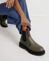 Bota tipo Chelsea de caña media, confeccionada en una combinación de cuero texturizado y gamuza en tonos verde oliva. Cuenta con paneles elásticos laterales en color negro, suela track de goma negra y tiradores en la parte superior en color bordó.