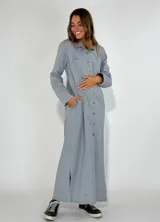 Vestido largo tipo camisa en color gris claro, con cuello camisero, botones frontales, mangas largas con puños abotonados, bolsillos de parche en el pecho y bolsillos laterales. Presenta aberturas laterales en el ruedo.