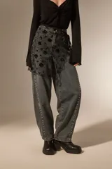 Falda mini de tiro alto confeccionada en denim gris oscuro con efecto lavado. Presenta un diseño superpuesto de tul negro con bordados florales y lentejuelas, además de detalles de tachas metálicas a lo largo de las costuras laterales.