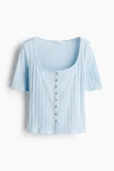 Top de manga corta de punto pointelle suave, color celeste, con ajuste entallado, escote en U y hilera de botones en la parte delantera. Presenta ribete levemente festoneado en el escote y la tapeta.