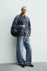 Bolso de hombro de diseño curvo en color negro, con detalle de pespuntes centrales. Cuenta con un compartimento principal con cierre de cremallera y dos bolsillos frontales con cierre. Incluye asa de hombro y bolsillo interior con cierre.