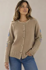 Saco de punto color beige, confeccionado en algodón, con cuello redondo, mangas largas y cierre frontal con botones. Presenta detalles bordados con diseño floral en las mangas y ribetes con rayas azules y blancas en la botonera y el cuello.