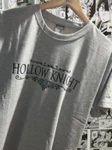 Remera gris de algodón con estampado frontal en negro del logo del videojuego "Hollow Knight", con tipografía decorativa y un emblema central.