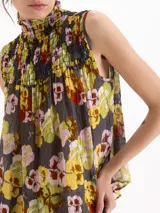 Top sin mangas confeccionado en crepé estampado con diseño floral en tonos amarillos, rosas y verdes sobre base gris. Cuenta con cuello alto fruncido y detalles de nido de abeja en el canesú.
