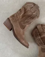 Bota corta de gamuza color marrón topo, estilo western, con bordados decorativos en hilo blanco en caña y empeine. Posee taco cuadrado de madera y punta ligeramente afilada.
