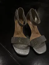 Sandalia de fiesta con taco cuadrado bajo, confeccionada en material plateado con acabado brillante. Presenta una tira frontal ancha y una pulsera al tobillo, ambas completamente cubiertas de strass.