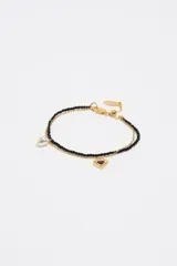 Pulsera doble dorada que combina una cadena de eslabones con una tira de cuentas negras de cristal. Cuenta con un dije colgante en forma de corazón esmaltado y un dije de perla barroca.
