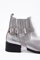 Botín de cuero vacuno en color plateado metalizado, con diseño de caña baja, elásticos laterales y detalle de estribo desmontable con hebilla metálica. Cuenta con un taco cuadrado bajo y suela negra.