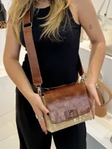 Bandolera de cuero genuino color marrón con solapa frontal y cuerpo confeccionado en cuero con pelo en tono dorado. Incluye una correa ancha de cuero regulable y extraíble con herrajes metálicos. El diseño presenta el logo de la marca grabado en un aplique de cuero sobre la solapa.