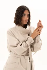 Trench corto color beige con cuello alto, cierre frontal de botones y cinturón ajustable en la cintura. Presenta mangas largas con detalles de tiras ajustables en los puños y bolsillos laterales.