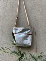 Cartera bandolera de cuero plateado con diseño flexible, trenza artesanal marrón al frente y correa larga ajustable con herrajes metálicos.