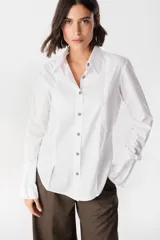 Camisa blanca de algodón con cuello clásico y cierre frontal de botones. Presenta un diseño con tablas verticales en el frente, corte redondeado en los laterales y puños anchos desmontables.