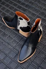 Bota corta de cuero negro con panel trasero de cuero vacuno blanco con manchas negras. Tiene cierre lateral y taco de madera.