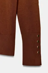 Jersey de punto liso color negro, con cuello redondo y manga larga. Presenta una hilera de cuatro botones dorados decorativos en el puño de cada manga.