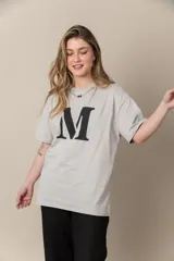 Remera gris con estampa de la letra M en negro y cadena plateada en el cuello.