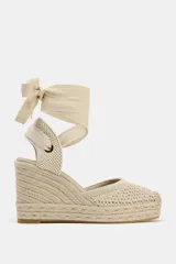Alpargata de mujer con plataforma y taco cuña alto, confeccionada en rafia trenzada. La capellada es de tejido de punto calado color beige, con talón cerrado y tiras largas de tela para atar al tobillo.