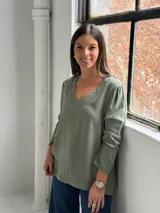 Blusa de manga larga en color verde oliva, con cuello en V y costuras decorativas visibles. Presenta un corte recto y holgado con un ligero fruncido en los hombros.