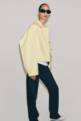 Chaqueta corta confeccionada en mezcla de algodón color amarillo claro, con cuello de solapa, manga larga, bolsillos delanteros y cierre frontal con botones visibles en los extremos.
