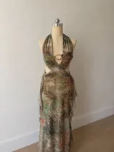 Vestido midi con estampado botánico en tonos tierra y verdes, con escote halter profundo y detalle de argolla metálica dorada en el centro. Presenta aberturas laterales en la cintura y falda con volados laterales.