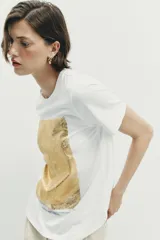 Camiseta de algodón con cuello redondo y manga corta. Presenta un diseño de estampación metalizada en color dorado en la parte frontal con detalles de texto.