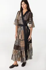 Vestido largo de algodón con patrón de rayas verticales y bloques de colores en tonos marrón, negro y crudo. Presenta diseño cruzado tipo envolvente con solapas negras, mangas tres cuartos acampanadas con volados, falda escalonada y lazo negro ajustable en la cintura.