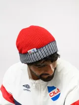 Gorro de lana tejido en color rojo con vuelta en azul marino y blanco a rayas. Incluye etiqueta lateral con el logo de Umbro y el escudo del Club Nacional de Football.